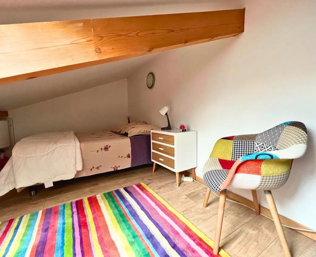 La chambre en mezzanine sous le toit, tapis à rayures multicolores et fauteuil patchwork